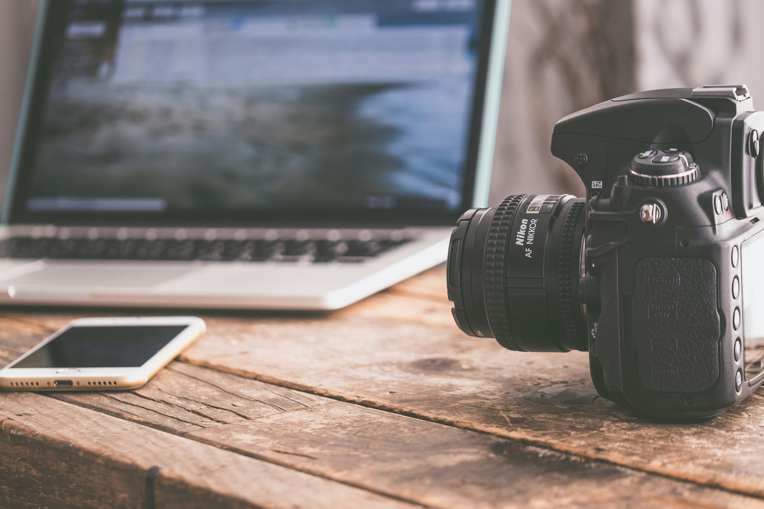 Close-up of a DSLR camera, laptop, and smartphone on a wooden table in a modern workspace.